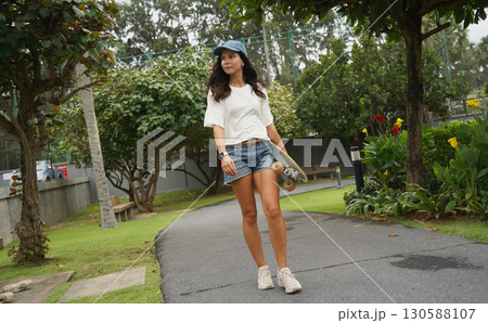 A woman is walking at the city park while carrying a skateboard in her hands 130588107