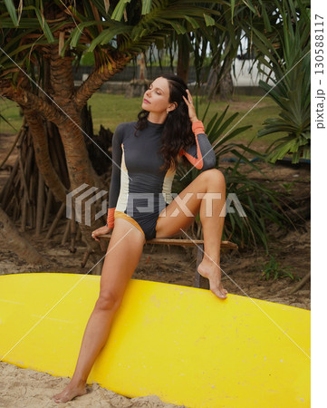 Woman in stylish wetsuit posing confidently with surfboard on beach Woman in stylish wetsuit posing confidently with surfboard on beach 130588117