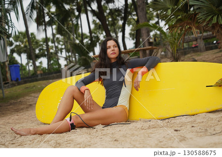 Woman in stylish wetsuit posing confidently with surfboard on beach 130588675