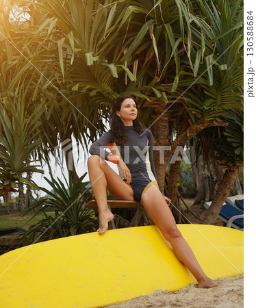 Woman in stylish wetsuit posing confidently with surfboard on beach Woman in stylish wetsuit posing confidently with surfboard on beach 130588684