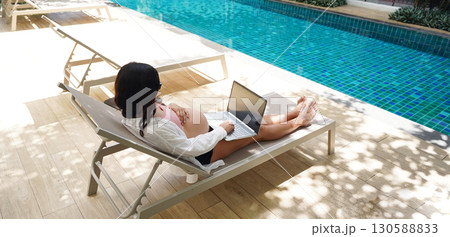 A pregnant woman is comfortably seated on a lounge chair using her laptop 130588833