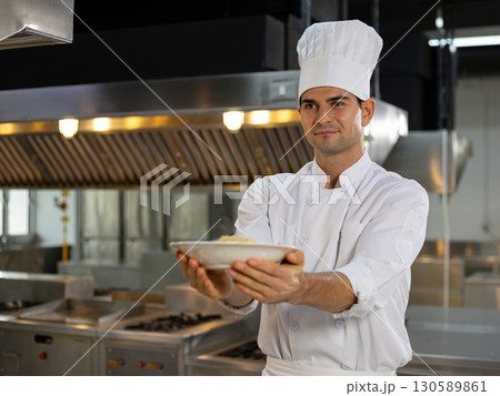 Smiling chef in uniform and hat proudly presents a finished dish while standing inside a modern stainless steel restaurant kitchen 130589861