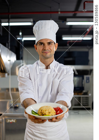 Professional chef in white uniform and hat proudly presenting a dish of fried rice with fresh vegetables inside a commercial kitchen 130589866