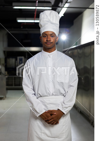 Confident young chef in white uniform and hat standing in a commercial kitchen, ready for cooking and culinary preparation 130589872