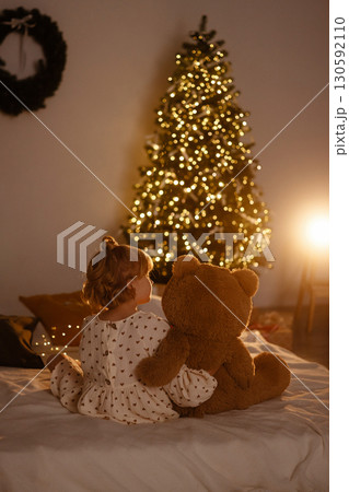 Girl sitting with teddy bear on bed rear view looking at Christmas tree with lights in cozy room 130592110