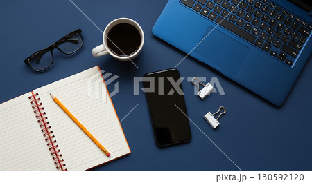 Minimalist flat lay of a blue laptop, open notebook, smartphone, coffee, and office supplies on a dark blue background, symbolizing organized work, study, and technology, perfect for productivity blog 130592120