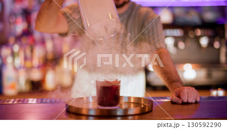 Bartender show. Drink presentation. Male barman lifting glass cylinder hood from smoke red alcoholic cocktail. Cool steam pub technique. 130592290