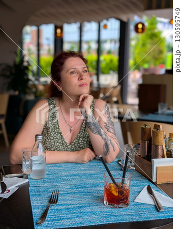 A woman sitting at a restaurant table with a cocktail. Lifestyle, dining, creativity, and relaxed urban atmosphere. A woman sitting at a restaurant table with a cocktail. Lifestyle, dining, creativity, and relaxed urban atmosphere. 130595749