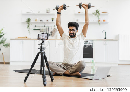 Male adult records fitness video blog using smartphone while using dumbbells for strength exercises. Seen seated in modern home interior, showing a health-focused and technology-driven lifestyle. 130595909