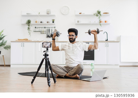 Man exercising with dumbbells during video recording in kitchen, showcasing fitness routine and health. Seated on mat near laptop and phone, promoting home fitness motivation. Man exercising with dumbbells during video recording in kitchen, showcasing fitness routine and health. Seated on mat near laptop and phone, promoting home fitness motivation. 130595936