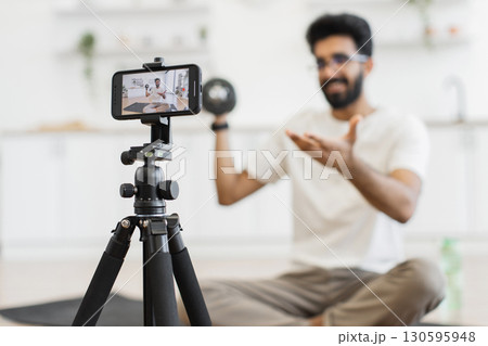 Young man with beard recording fitness video at home, sitting on floor describing techniques for exercising with dumbbells. View shows person filming on smartphone positioned on tripod Young man with beard recording fitness video at home, sitting on floor describing techniques for exercising with dumbbells. View shows person filming on smartphone positioned on tripod 130595948