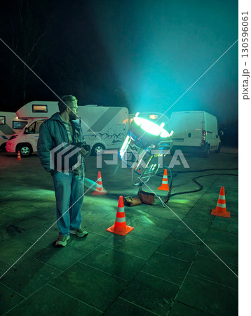 Man standing near cinema spotlight on outdoor film set at night. Movie production, technology, and artistic atmosphere in cinematography. 130596061