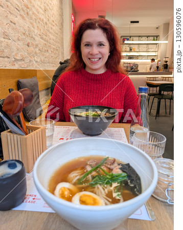 A woman in a red sweater smiles at a table with ramen bowls inside a restaurant. Dining culture, lifestyle, gastronomy and modern culinary tradition in urban hospitality. A woman in a red sweater smiles at a table with ramen bowls inside a restaurant. Dining culture, lifestyle, gastronomy and modern culinary tradition in urban hospitality. 130596076