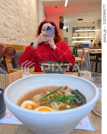 A woman in a red sweater takes a photo of ramen bowls at a restaurant table. Dining culture, lifestyle, gastronomy and modern food photography in casual urban hospitality. 130596077