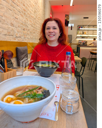 A woman in a red sweater smiles at a table with ramen bowls inside a restaurant. Dining culture, lifestyle, gastronomy and modern culinary tradition in urban hospitality. A woman in a red sweater smiles at a table with ramen bowls inside a restaurant. Dining culture, lifestyle, gastronomy and modern culinary tradition in urban hospitality. 130596079
