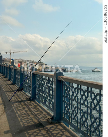 Fishing rod leaning on bridge railing with sea and boats in background. Urban lifestyle, leisure, and traditional activity in city environment. Fishing rod leaning on bridge railing with sea and boats in background. Urban lifestyle, leisure, and traditional activity in city environment. 130596255