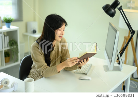 Young woman working at home office desk while reading notes from a journal Young woman working at home office desk while reading notes from a journal 130599831