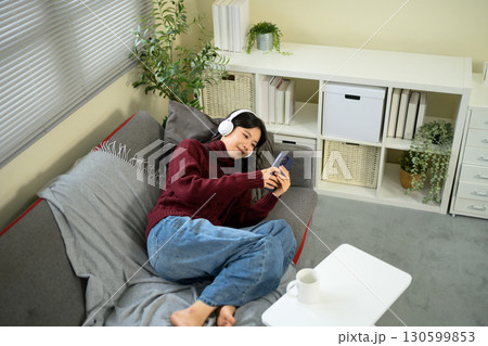 Young woman relaxing on a sofa at home, wearing headphones and using smartphone 130599853