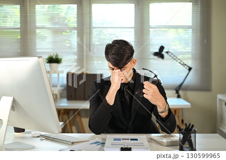 Stressed businessman holding glasses and rubbing eyes while working at desk in modern office Stressed businessman holding glasses and rubbing eyes while working at desk in modern office 130599965
