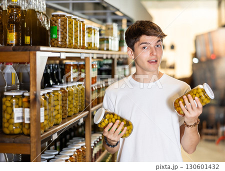Interested guy choosing pickled olives in glass jars in supermarket Interested guy choosing pickled olives in glass jars in supermarket 130601432