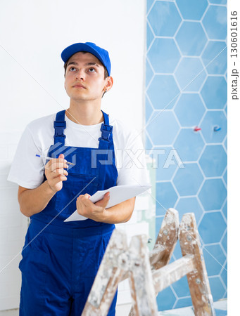 Portrait of man builder in construction overalls with documents in hands checks quality of completed repairs in apartment Portrait of man builder in construction overalls with documents in hands checks quality of completed repairs in apartment 130601616