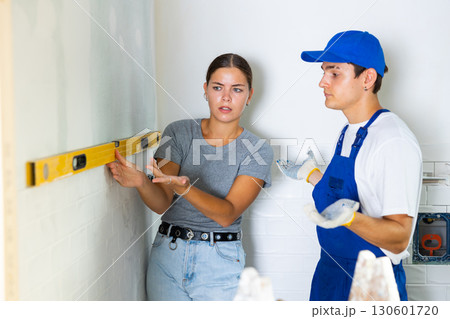 Young woman uses a spirit level on wall, discussing and showing something to worker Young woman uses a spirit level on wall, discussing and showing something to worker 130601720