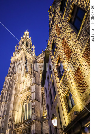 Looking up at the illuminated Cathedral of Our Lady in Antwerp, Belgium, at dusk, with the intricate Gothic architecture towering against the deep blue sky next to a beautifully textured building Looking up at the illuminated Cathedral of Our Lady in Antwerp, Belgium, at dusk, with the intricate Gothic architecture towering against the deep blue sky next to a beautifully textured building 130602480