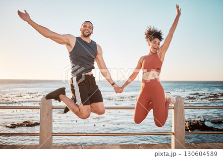 Fitness, energy and portrait of a couple jumping at beach for training fun, support and celebration of goal. Jump, happy and excited man and woman holding hands at the sea for cardio in Spain 130604589