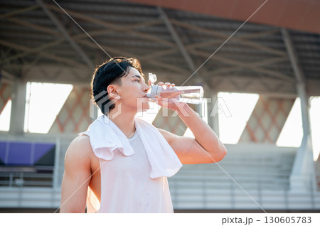 Asian man in white tank top standing and drinking from a water bottle, taking a rest after running. 130605783