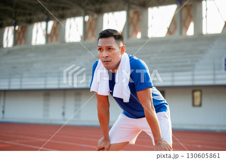 Asian man in blue t shirt standing with hands on his knee, taking a rest after running on racetrack. Asian man in blue t shirt standing with hands on his knee, taking a rest after running on racetrack. 130605861