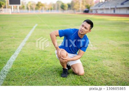 Asian man in blue shirt kneeling on grass and holding his knee while looking up. Leg pain or injury. 130605865