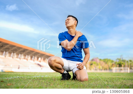 Asian man in blue shirt is kneeling on grass and touching or massaging his shoulder. Pain or injury. 130605868