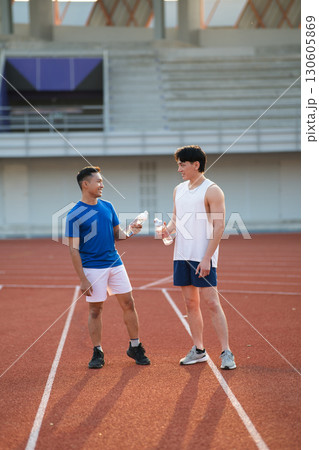 Two asian athlete men smiling at each other while holding water bottles and standing on racetrack 130605869
