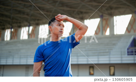 Asian man in blue t shirt is standing and wiping his sweat with a hand, taking a rest after running. Asian man in blue t shirt is standing and wiping his sweat with a hand, taking a rest after running. 130605905