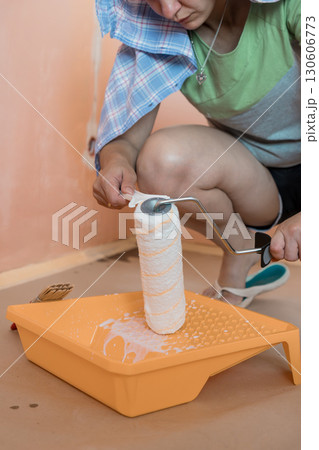 Young Woman Painting a Peach-Colored Wall Indoors with Roller, Paint Tray, and Checkered Shirt, Home Renovation Scene 130606773