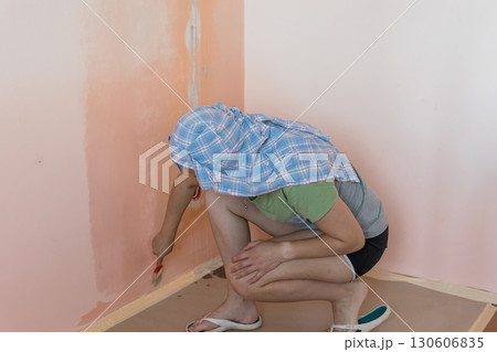 Home Renovation Concept: Woman Painting Peach-Colored Wall in Room Corner with Blue Shirt and Painters Tape. 130606835