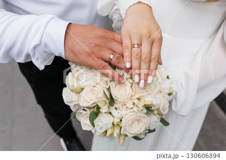 Wedding Rings on Hands with Bouquet, Marriage Symbolism, White Roses, Elegant Ceremony, Love, Commitment, Romance, Neutral Tones, Close-Up Shot 130606894
