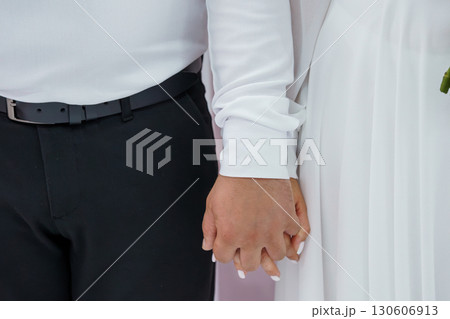 Close-up of Couple Holding Hands, Wedding Day Romance, Black Suit, White Dress, Celebration, Love, Formal Attire, Neutral Background, Stylish. Close-up of Couple Holding Hands, Wedding Day Romance, Black Suit, White Dress, Celebration, Love, Formal Attire, Neutral Background, Stylish. 130606913