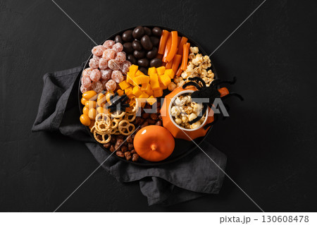 Halloween sweet charcuterie board with pumpkin, popcorn, color chocolate candy, spider, crackers on black background. View from above. Halloween sweet charcuterie board with pumpkin, popcorn, color chocolate candy, spider, crackers on black background. View from above. 130608478