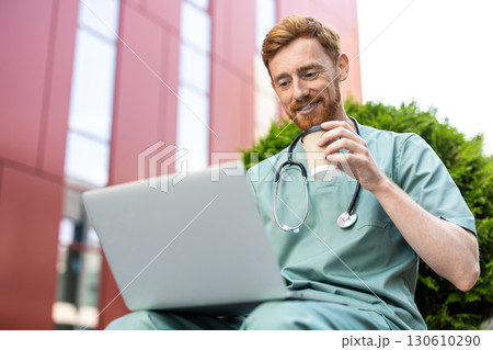 Ginger man doctor in scrubs enjoying break with laptop and hot drink 130610290