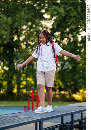 Girl with a red backpack walking on the log on a playground and keeping balance Girl with a red backpack walking on the log on a playground and keeping balance 130610402