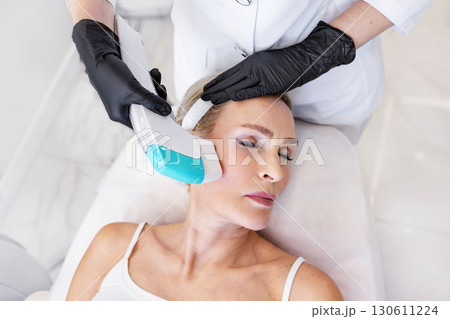 Cosmetologist in black gloves performing hardware facial treatment with modern device on mature woman lying in clinic. Concept of beauty, anti-aging, dermatology, skincare, rejuvenation 130611224