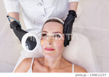 Cosmetologist performing photorejuvenation facial treatment with modern equipment for mature woman, lying on chair during non-invasive anti-aging skincare procedure in clean luxury beauty clinic 130611237