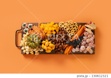 Halloween sweet charcuterie board with pumpkin, popcorn, dried fruits, color chocolate candy, spider, crackers on cutting board on orange background. Festive food for kids party. View from above. Halloween sweet charcuterie board with pumpkin, popcorn, dried fruits, color chocolate candy, spider, crackers on cutting board on orange background. Festive food for kids party. View from above. 130612321