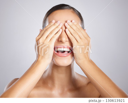 Woman, hands and covering eyes with smile in satisfaction for skincare, cosmetics or beauty against a grey studio background. Happy female model smiling with covered face for makeup surprise 130613921