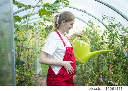 Pregnant woman watering plants in greenhouse with watering can Pregnant woman watering plants in greenhouse with watering can 130614314