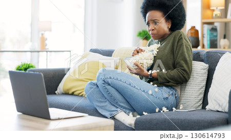 African woman, watching and horror movie on a laptop eating popcorn on a living room couch. Scared female person, home lounge and online series with film snack and food with scary video on computer 130614793