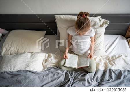 Little girl sitting on her parents bed and reading a book in the bedroom. Concept of quiet time, concentration and love for reading in early childhood. Little girl sitting on her parents bed and reading a book in the bedroom. Concept of quiet time, concentration and love for reading in early childhood. 130615082