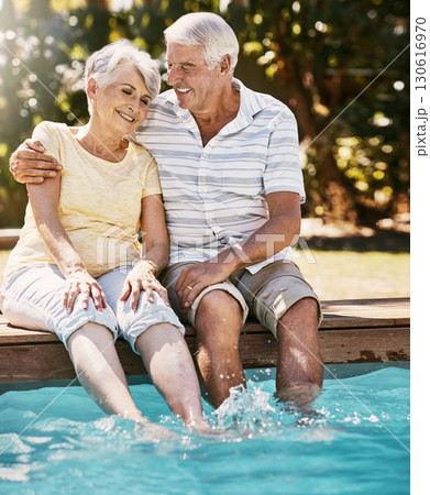 Elderly couple, hug and smile by swimming pool for relax, love or quality bonding time together on summer vacation. Happy senior man holding woman relaxing with feet in water by the poolside outside 130616970