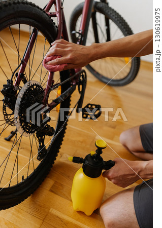 Close-up of cyclist male cleaning dirty mountain bike indoors using water sprayer and cloth to maintain equipment after exhilarating ride, ensuring optimal performance. Concept of bicycle service. 130619975